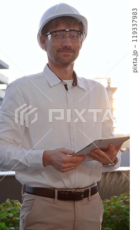 Man an architectural engineer, wearing a white shirt and hard hat, is keeping digital tablet and looking at the camera while at a building site at sunrise, front and vertical view Man an architectural engineer, wearing a white shirt and hard hat, is keeping digital tablet and looking at the camera while at a building site at sunrise, front and vertical view 119337983