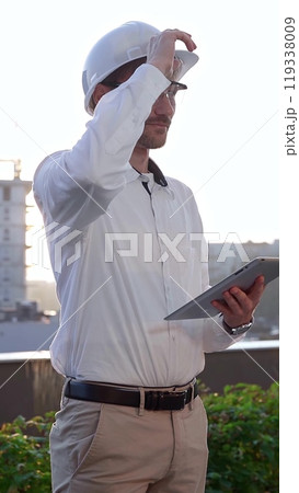 Male architectural engineer, wearing a white shirt and hard hat, is using a digital tablet while inspecting a building site at sunrise, front and vertical view. Construction and engineering Male architectural engineer, wearing a white shirt and hard hat, is using a digital tablet while inspecting a building site at sunrise, front and vertical view. Construction and engineering 119338009