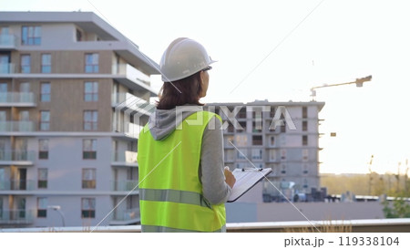 Female engineer with a white protective helmet and safety vest is writing on a clipboard while inspecting a construction site at sunset. Architect concept 119338104