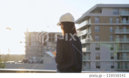 Female architect wearing a safety helmet is using a tablet computer while inspecting a building construction site at sunset 119338141