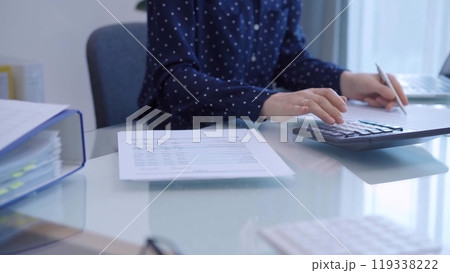 A female accountant with blue dotted blousy is using a laptop computer and calculator to calculate taxes at a glass desk in the office 119338222