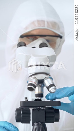 Scientist in ppe analyzing samples with microscope. Close-up of a focused researcher in a lab coat and protective gear using a microscope in a laboratory setting Scientist in ppe analyzing samples with microscope. Close-up of a focused researcher in a lab coat and protective gear using a microscope in a laboratory setting 119338229