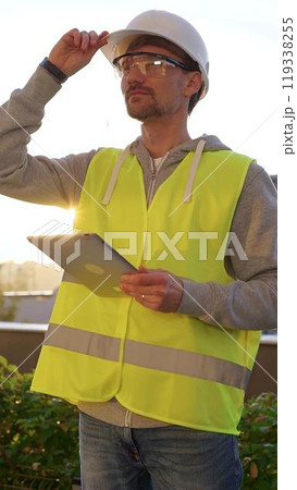 Man engineer with white helmet and safety vest is taking notes on a digital tablet while inspecting a construction site at sunrise in early morning, vertical front view. Architecture concept Man engineer with white helmet and safety vest is taking notes on a digital tablet while inspecting a construction site at sunrise in early morning, vertical front view. Architecture concept 119338255