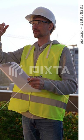 Man engineer with white helmet and safety vest is taking notes on a clipboard while inspecting a construction site at sunrise in early morning, vertical front view. Architecture and engineering Man engineer with white helmet and safety vest is taking notes on a clipboard while inspecting a construction site at sunrise in early morning, vertical front view. Architecture and engineering 119338281