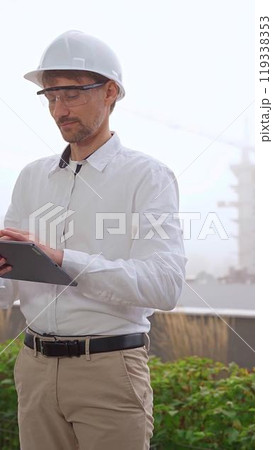 Male construction engineer or architect, wearing a white shirt and helmet, is using a digital tablet while inspecting a building site in foggy weather, front view. Architecture concept Male construction engineer or architect, wearing a white shirt and helmet, is using a digital tablet while inspecting a building site in foggy weather, front view. Architecture concept 119338353