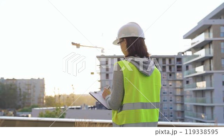 Female engineer with a white protective helmet and safety vest is writing on a clipboard while inspecting a construction site at sunset. Architect and engineering concept Female engineer with a white protective helmet and safety vest is writing on a clipboard while inspecting a construction site at sunset. Architect and engineering concept 119338595