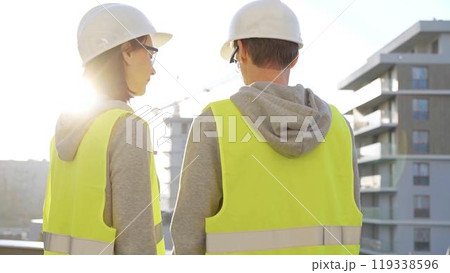 Man and woman engineers wearing safety hard hats and vests holding blueprint and discussing something on construction site at sunrise, back view. Architecture and engineering concept Man and woman engineers wearing safety hard hats and vests holding blueprint and discussing something on construction site at sunrise, back view. Architecture and engineering concept 119338596