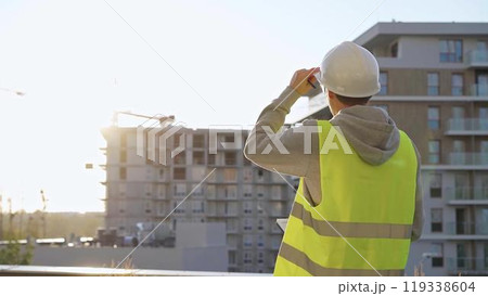 Man engineer with white helmet and safety vest is taking notes with a tablet computer while inspecting a construction site at sunrise 119338604