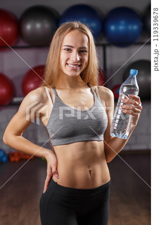 Young woman holding a water bottle and smiling in a gym during fitness session 119339678