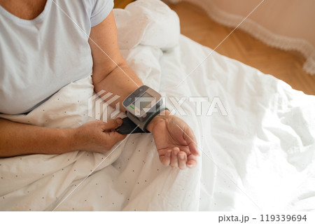 Close up of woman checks her blood pressure, a tonometer is worn on her wrist in a bed. Medical device for measuring blood pressure and heart rate used at hand wrist 119339694