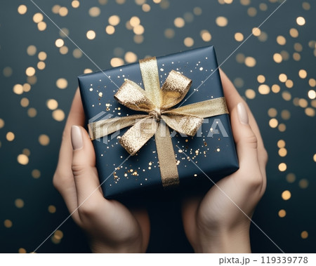 A person holding a beautifully wrapped gift with a gold bow, surrounded by a dreamy bokeh of golden lights, perfect for celebrations and special moments. 119339778