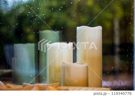 Set of white yellow paraffin artificial tall candles against wet window with water droplets, fall rain outside a window Decor for cozy home comfort. Autumnal cold rainy weather still life. Fall season 119340776