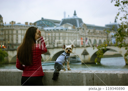 A beautiful young woman in red jacket with small Yorkshire Terrier dog sitting on parapet standing on the banks of a River Seine in Paris. Journey to France with pet. Walking outside on an autumn day. A beautiful young woman in red jacket with small Yorkshire Terrier dog sitting on parapet standing on the banks of a River Seine in Paris. Journey to France with pet. Walking outside on an autumn day. 119340890