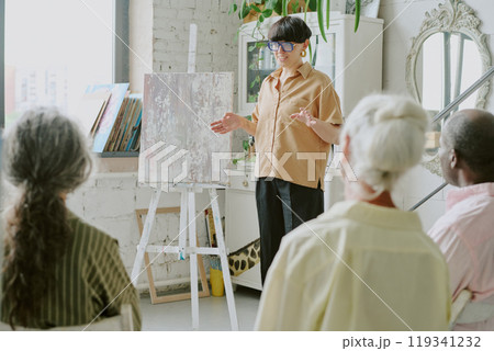 Talented young Caucasian teacher standing next to white wooden easel while unrecognizable senior students listening to her Talented young Caucasian teacher standing next to white wooden easel while unrecognizable senior students listening to her 119341232