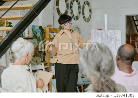 Young confident teacher holding huge canvas while explaining drawing tips to senior students 119341305