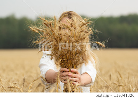 Female farmer is holding a big bundle of wheat ears in front of her in the golden agricultural field, forest in the background. 119341320