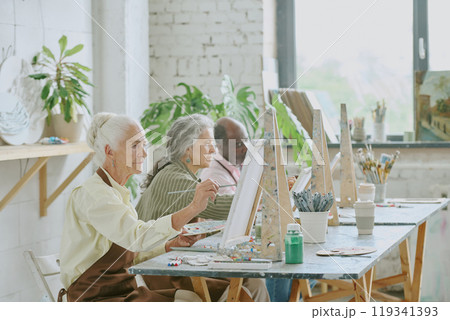 Senior biracial people sitting at dirty wooden table with easel, canvas and painting tools while painting together Senior biracial people sitting at dirty wooden table with easel, canvas and painting tools while painting together 119341393