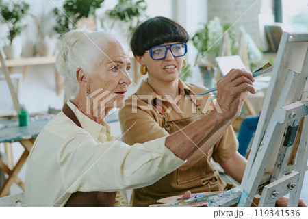 Mature drawing teacher with bob haircut helping her senior student who trying to paint on canvas with oil paints 119341536