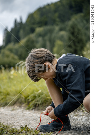 Young boy kneeling on a footpath in green nature to tie his hiking shoes Young boy kneeling on a footpath in green nature to tie his hiking shoes 119341543