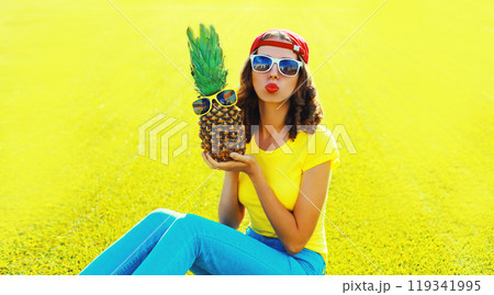 Summer portrait happy young woman blowing kiss with pineapple, glasses on the grass in summer park Summer portrait happy young woman blowing kiss with pineapple, glasses on the grass in summer park 119341995