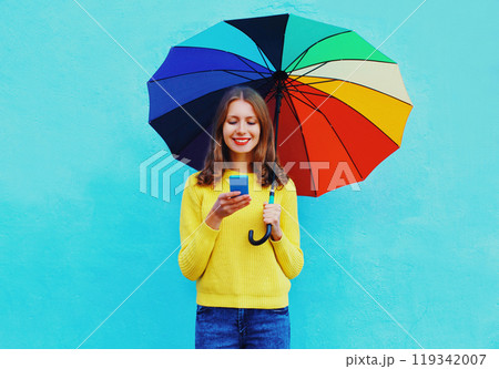 Happy smiling young woman looking at mobile phone with colorful umbrella on blue studio background 119342007