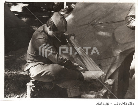 Photo of a young soldier in military uniform in front of the tent. The young man puts on footcloths. 119342288
