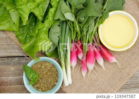 Fresh ripe vegetables on the wooden table with burlap table cloth - radish, green onion, lettuce, dried herbs in a little prep bowl with basil leaves and olive oil are ready to cook salad. Fresh ripe vegetables on the wooden table with burlap table cloth - radish, green onion, lettuce, dried herbs in a little prep bowl with basil leaves and olive oil are ready to cook salad. 119347387