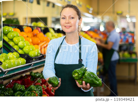 Professional adult female seller in apron putting red and green bell peppers on stalls in hypermarket 119349421