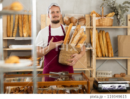 In sales area room of bakery, there is guy with loaf breads and baguetttes in wicker basket in hands In sales area room of bakery, there is guy with loaf breads and baguetttes in wicker basket in hands 119349615