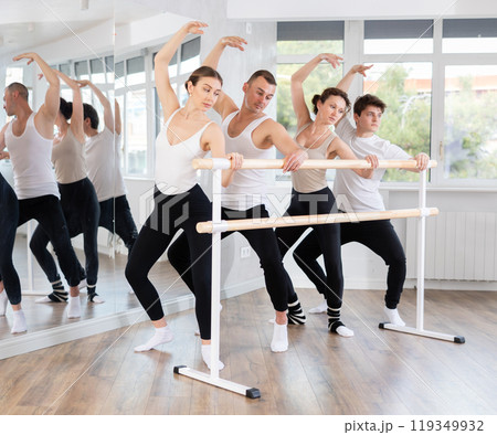 Group of sports people standing in row together while doing ballet dance moves demi plie while holding on ballet barre in ballet studio 119349932