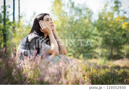 A young woman sits peacefully in a sunlit forest clearing, enjoying nature A young woman sits peacefully in a sunlit forest clearing, enjoying nature 119352930