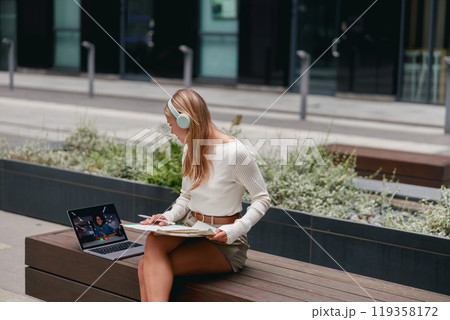 Stylish Young Woman Effectively Working Outdoors Using Her Laptop and Headphones 119358172