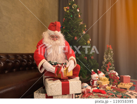 Santa Claus with gift box sitting on sofa at home near Christmas tree, presents, snowmen decorations and festive ornaments at night 119358395