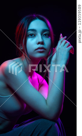 Portrait of beautiful young Korean girl posing against black background with blue and pink neon lights 119359589
