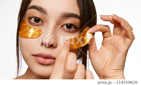 Korean woman applying golden under-eye patches, highlighting a skincare routine for reducing puffiness and brightening the under-eye area Korean woman applying golden under-eye patches, highlighting a skincare routine for reducing puffiness and brightening the under-eye area 119359620