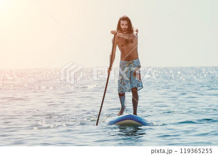 Man Paddleboarding Ocean Sunset - A man is paddleboarding in the ocean at sunset. Man Paddleboarding Ocean Sunset - A man is paddleboarding in the ocean at sunset. 119365255