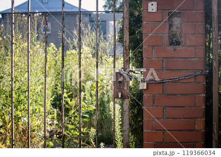 Rusty old lock hangs on a chain at the gate on a red brick post on abandoned territory with greenery 119366034