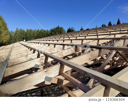 Side view of wooden roof trusses on a construction site with a bright blue sky, showing the intricate assembly and metal connectors Side view of wooden roof trusses on a construction site with a bright blue sky, showing the intricate assembly and metal connectors 119366304