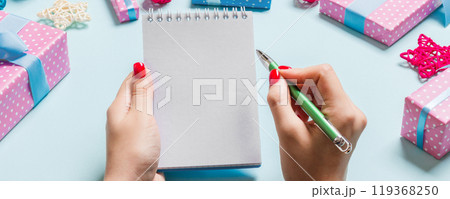 Top view of female hand making some notes in noteebok on blue background. New Year decorations and toys. Christmas time concept Top view of female hand making some notes in noteebok on blue background. New Year decorations and toys. Christmas time concept 119368250