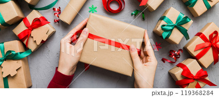 Woman is packing surprise gift for Christmas holidays, sitting at workplace with craft tools, top view. Woman wrapping Christmas gift at colored table 119368284