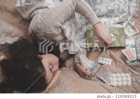 a sick girl in a sweater lying on the bed checks the temperature with a thermometer on the background of pills scattered on the blanket 119369223