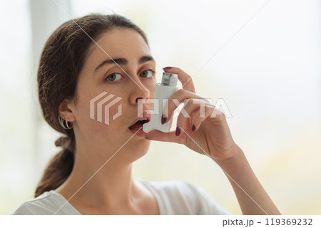 Asthma. Portrait of a young woman holding an inhaler to her mouth and looking at the camera. Blurry background. Copy space Asthma. Portrait of a young woman holding an inhaler to her mouth and looking at the camera. Blurry background. Copy space 119369232