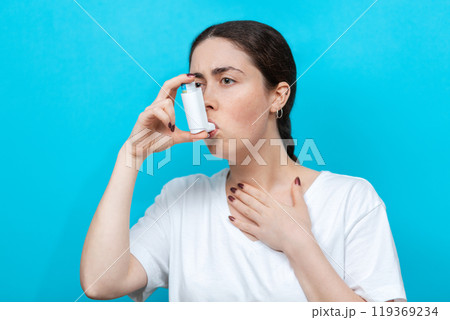 Asthma. Portrait of a young woman using an inhaler. Blue background Asthma. Portrait of a young woman using an inhaler. Blue background 119369234