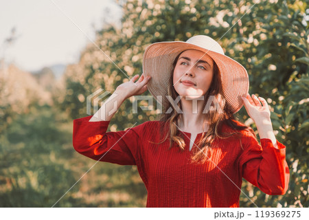 Portrait of a woman in the garden. A young and beautiful woman in a red dress holds her hat and looks dreamily into the distance Portrait of a woman in the garden. A young and beautiful woman in a red dress holds her hat and looks dreamily into the distance 119369275