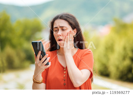Portrait of a woman looking at the phone, clutching her face in fear. The Park is in the background. Concept of online fraud and fake news 119369277