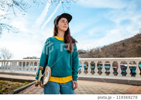 Portrait of a Young Caucasian woman holding a skateboard. In the background, the blue sky and the Boulevard. Copy space. Concept of sports lifestyle and street culture 119369278