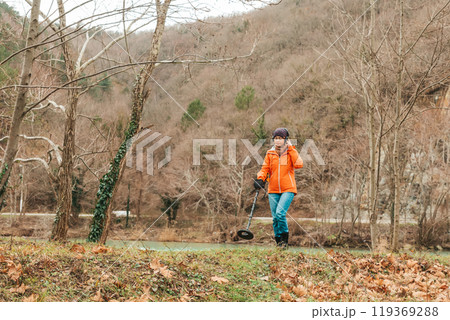Search With A Metal Detector. A young woman in an orange jacket is looking for lost artifacts in the Park area. Hobbies and activities Search With A Metal Detector. A young woman in an orange jacket is looking for lost artifacts in the Park area. Hobbies and activities 119369288