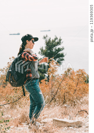 Tourism and outdoor activities. A woman in a cap and glasses, wearing a backpack. In the background the sea and trees. Vertical orientation 119369305