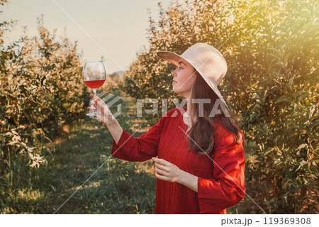 Wine tasting and relaxing in the garden. Young beautiful brunette in a red dress and hat holding a glass of wine. There are fruit trees in the background. Tint Wine tasting and relaxing in the garden. Young beautiful brunette in a red dress and hat holding a glass of wine. There are fruit trees in the background. Tint 119369308