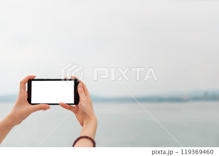 Mock up. Women's hands holding the phone horizontally. In the background sea and sky. Copy space. Isolate 119369460